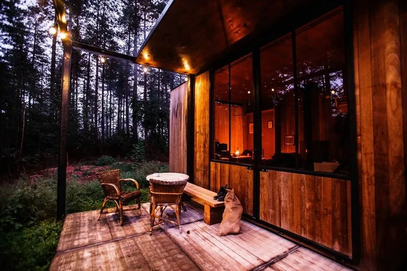 A cozy wooden cabin with large glass windows, surrounded by trees, features a small table, chairs, and a dog on the deck at dusk.