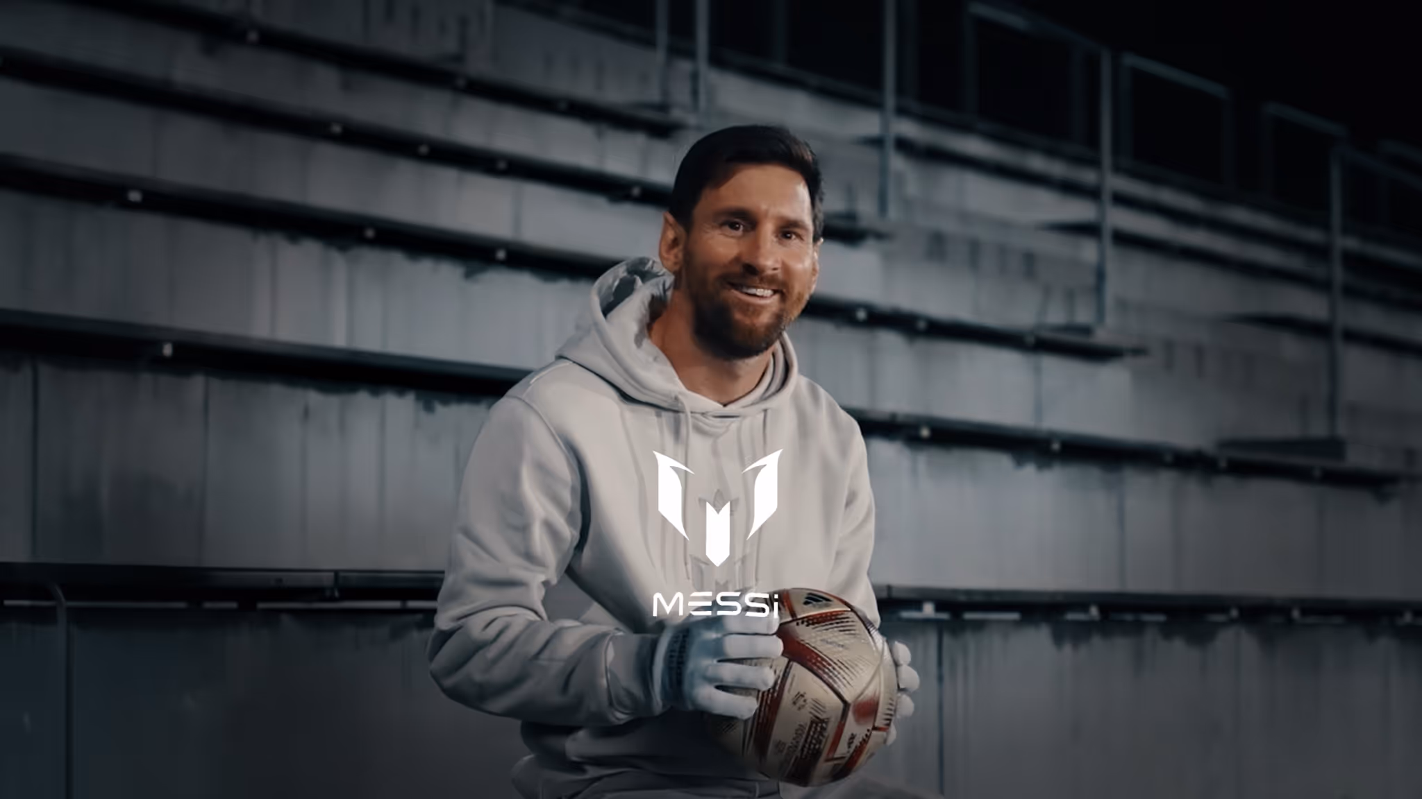 Lionel Messi in a light gray hoodie and gloves holding a soccer ball, sitting in an empty stadium with a Messi logo overlay.