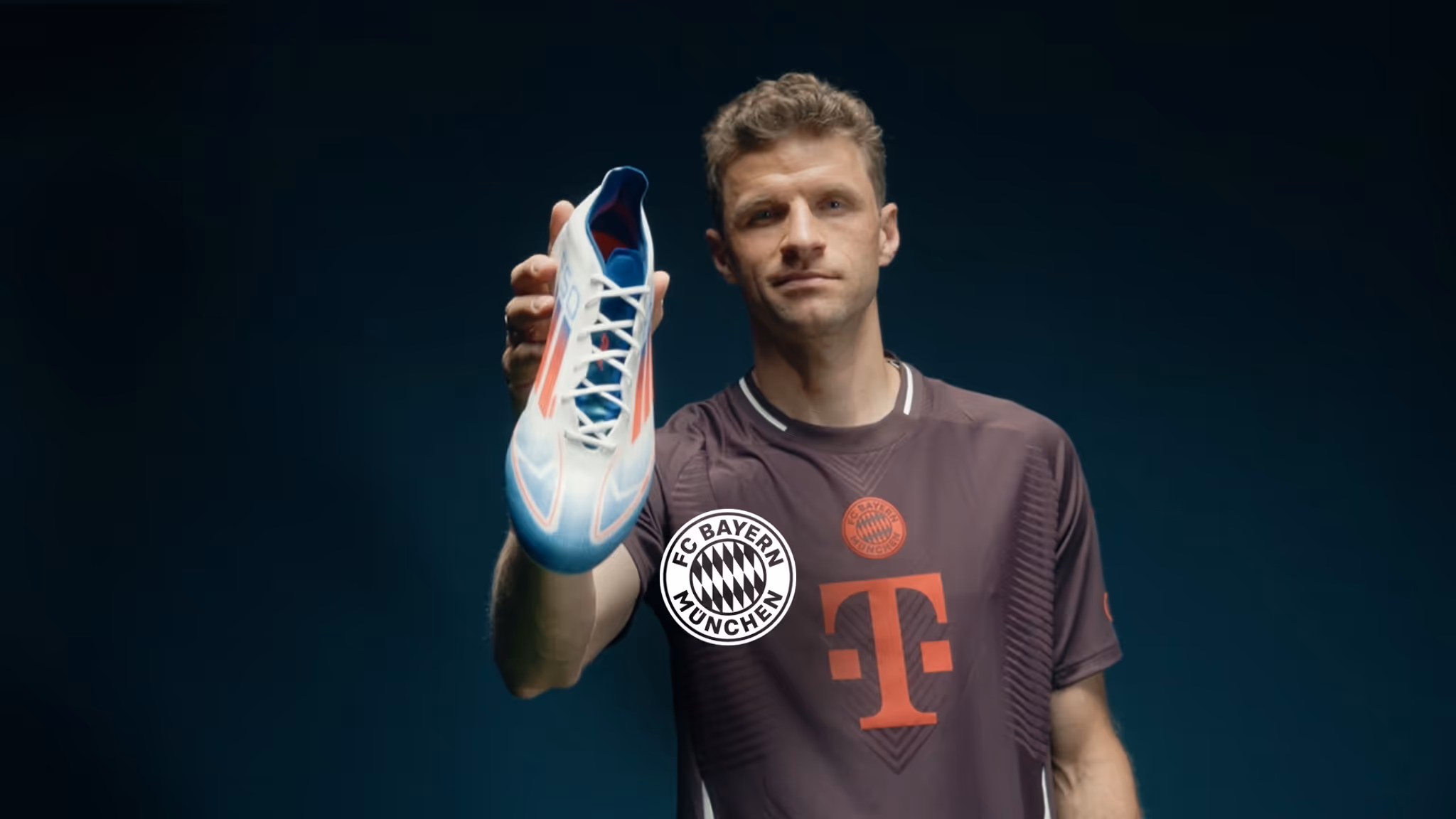 Thomas Müller wearing an FC Bayern München jersey holds a white and blue soccer shoe towards the camera.