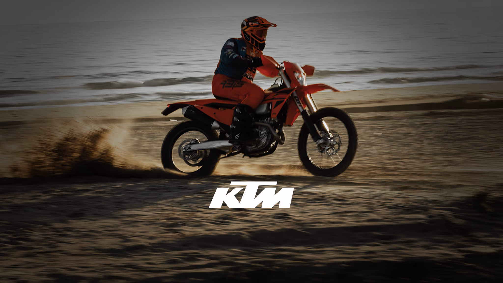 Motorcyclist in orange gear riding a dirt bike on sand near water with KTM logo overlaid.