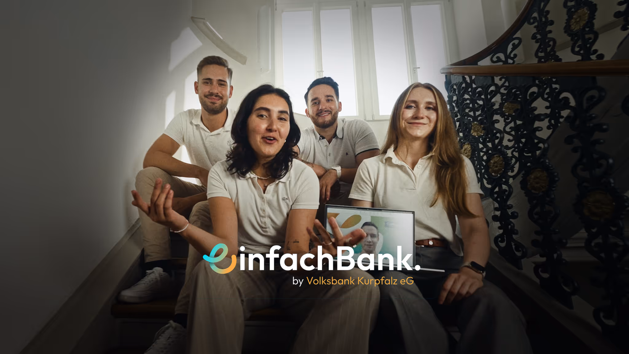 Four young adults sitting on stairs, smiling, with one holding a laptop showing a fifth person via video call.