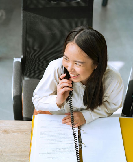 Professional woman smiling while talking on office phone with documents on desk
