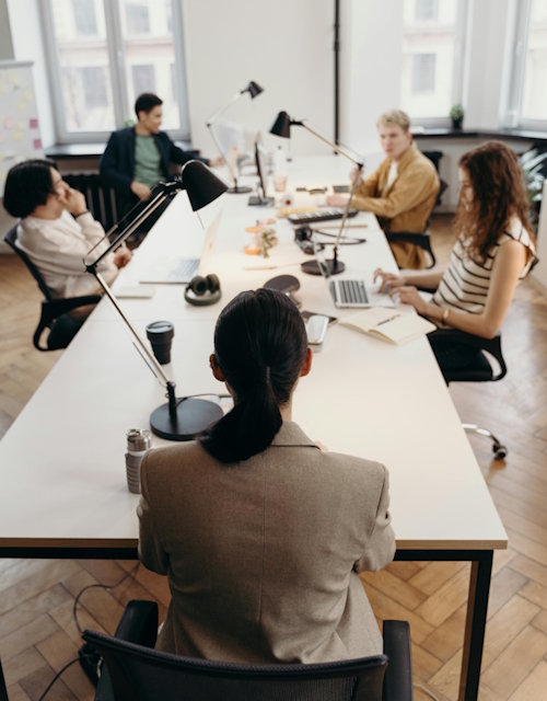 Team collaborating in a modern office, seated around a long shared desk with laptops and notebooks.