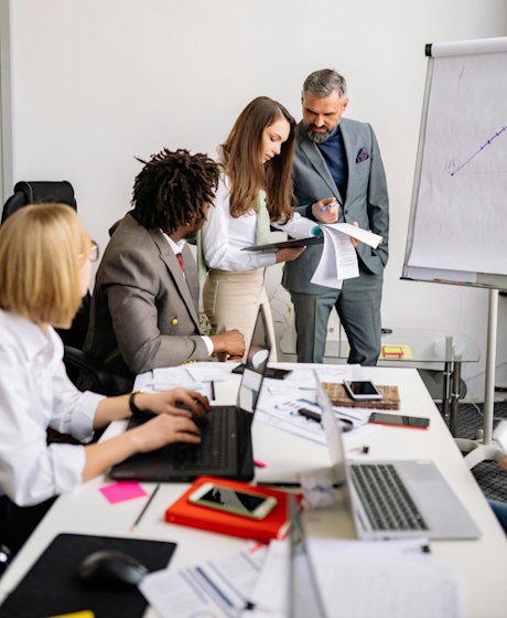 Team meeting with members reviewing reports and working on laptops around table with whiteboard in background