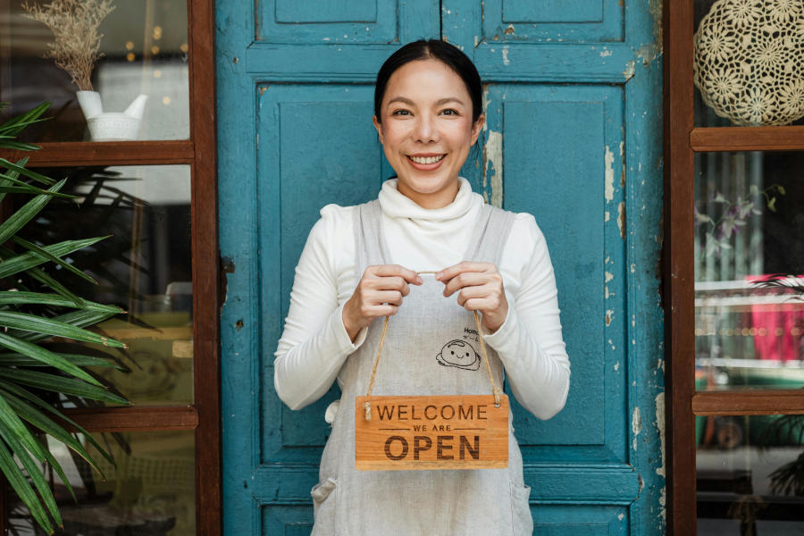 A small business owner in white clothing holds up a wooden "Welcome We Are Open" sign while smiling in front of a turquoise door.