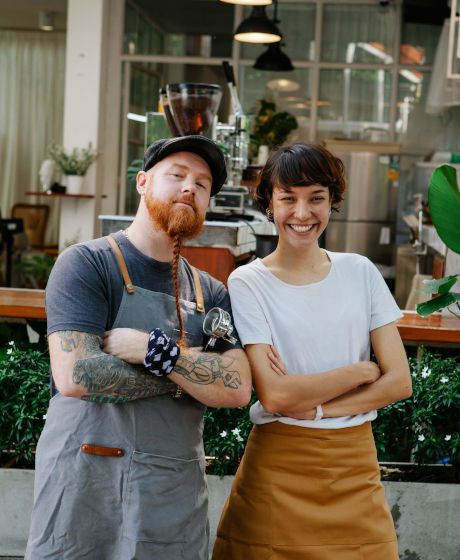 Two smiling workers stand together in a cafe, with one wearing an apron and hat.