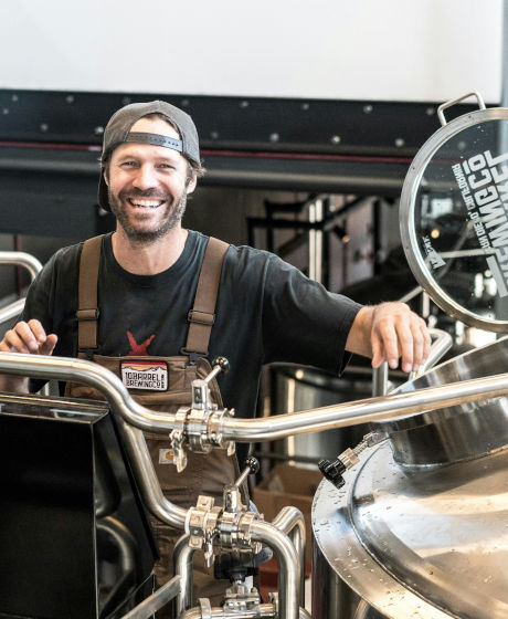 A man in a cap and apron smiles while standing at large metal brewing tanks in a brewery.
