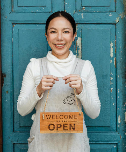 A small business owner in white clothing holds up a wooden "Welcome We Are Open" sign while smiling in front of a turquoise door.