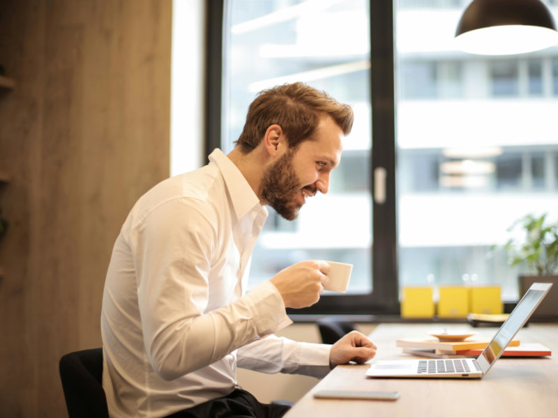 Smiling professional enjoying coffee while working on laptop at desk in bright modern office