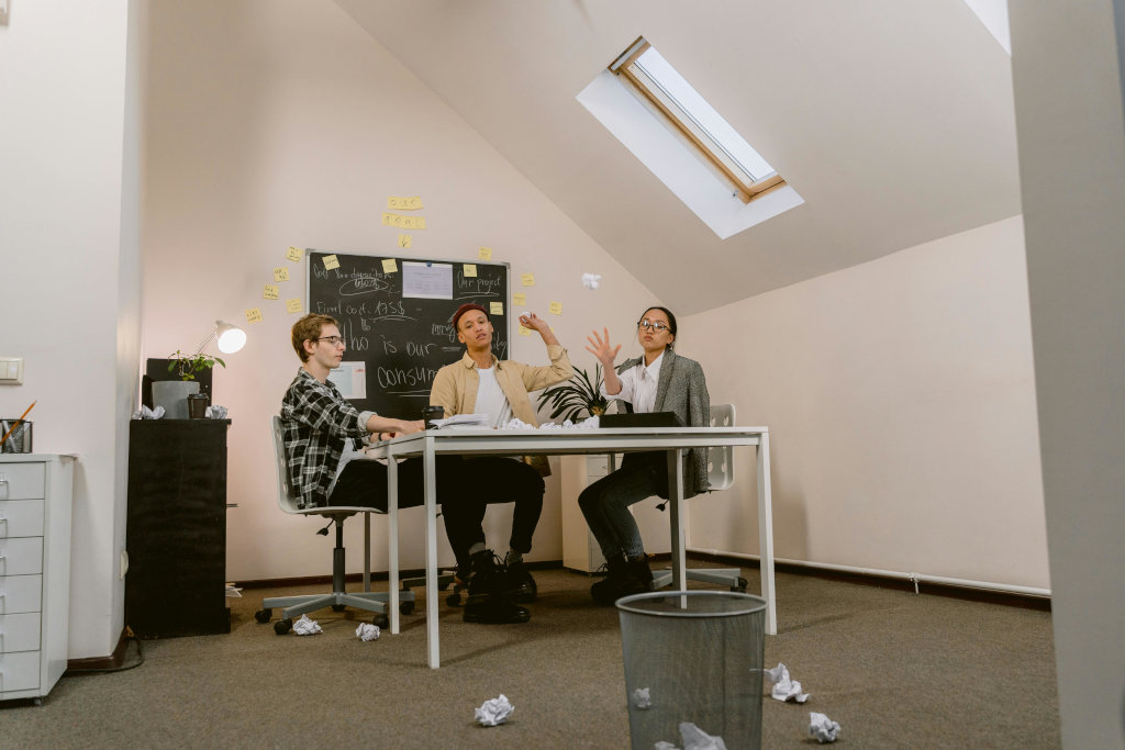 Three people sit at a table in a bright office with a chalkboard behind them, tossing crumpled papers into a bin