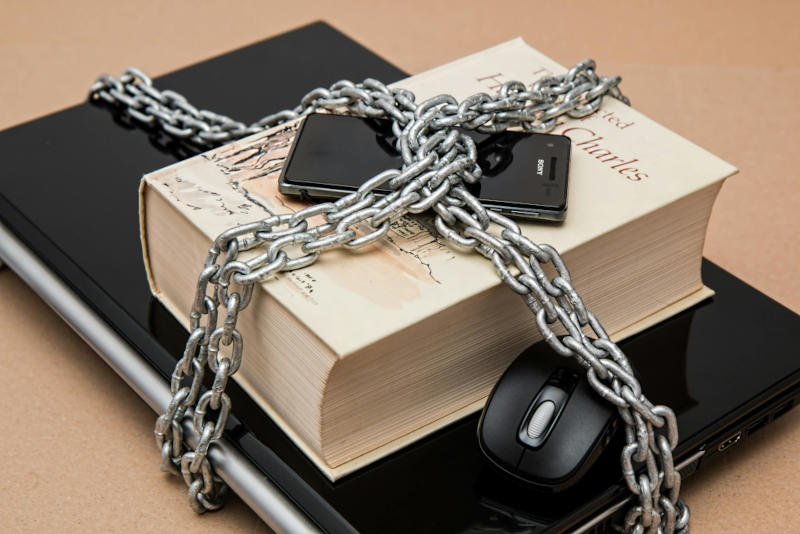 A thick metal chain wrapped around stacked books, a smartphone, and a computer mouse.