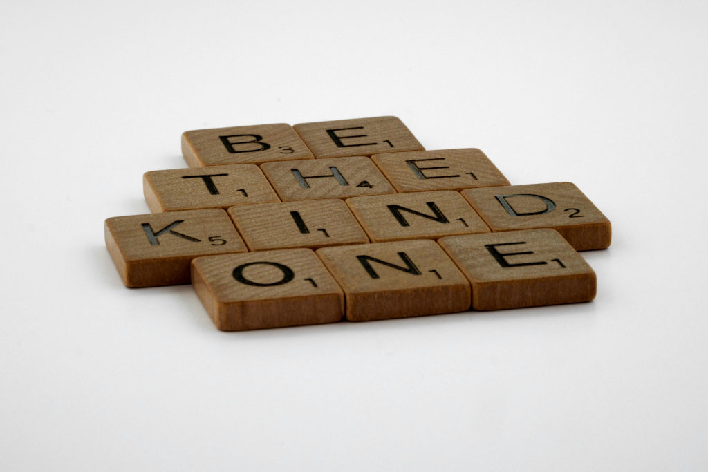 Wooden Scrabble tiles arranged to spell "Be the kind one" against a white background.