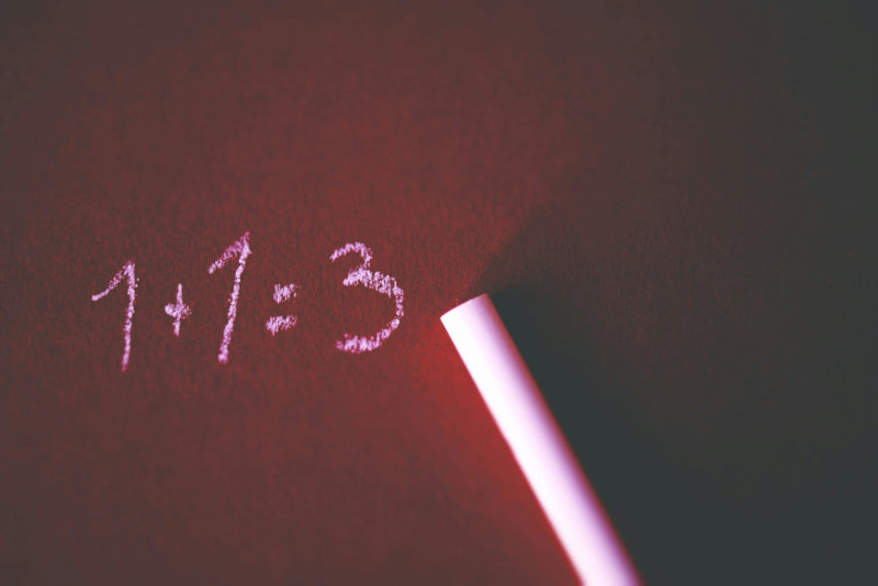 A piece of chalk lying on a dark red surface next to a handwritten equation ‘1 + 1 = 3’ written in pink chalk, illustrating a simple mathematical mistake.