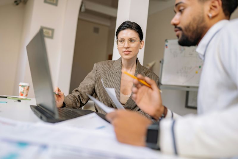 Two professionals collaborating at a desk with a laptop and documents in an office setting.