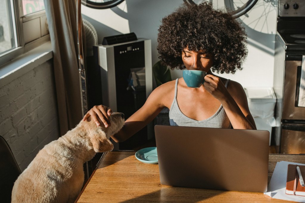 Woman working at a laptop while petting a small dog and drinking from a blue mug.