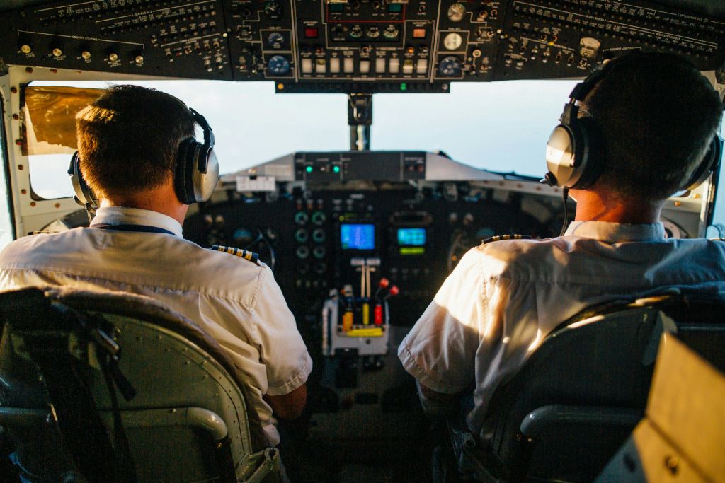 Two pilots in an aircraft cockpit wearing headsets, viewed from behind as they operate the controls.