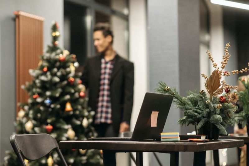 Office desk decorated for the holidays with a computer monitor and festive floral arrangement, with a decorated Christmas tree and person visible in the blurred background.