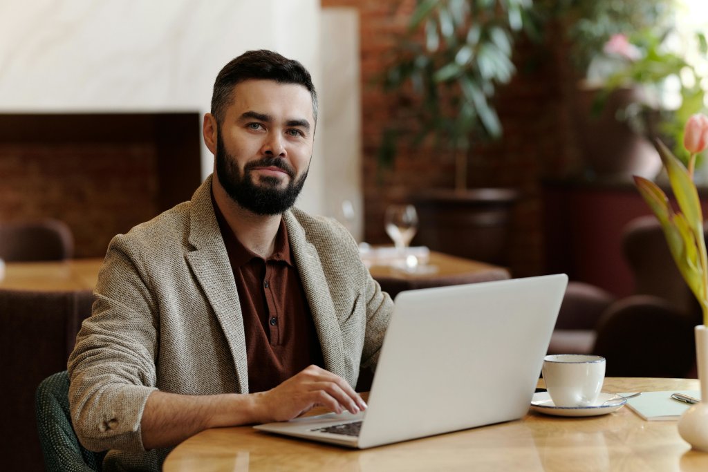 Man working on a laptop at a table in a modern office or café setting with plants and a coffee cup nearby.