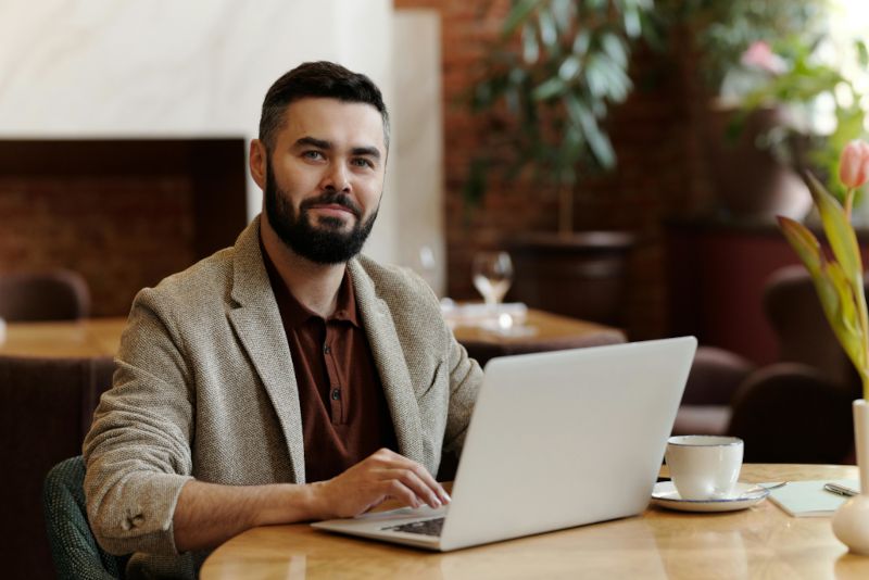 Man working on a laptop at a table in a modern office or café setting with plants and a coffee cup nearby.