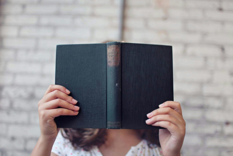 Person reading an open hardcover book with their face obscured, standing in front of a white brick wall.