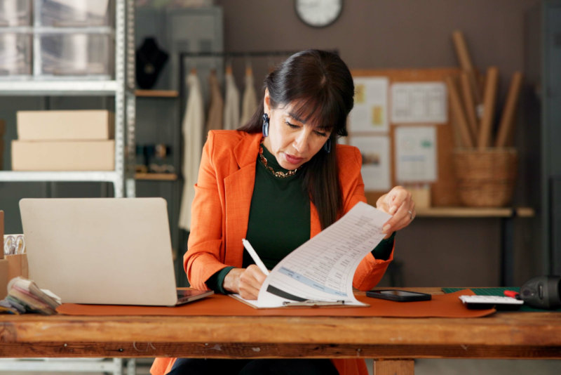 Woman in an orange blazer writing on a document while working at a desk with a laptop.