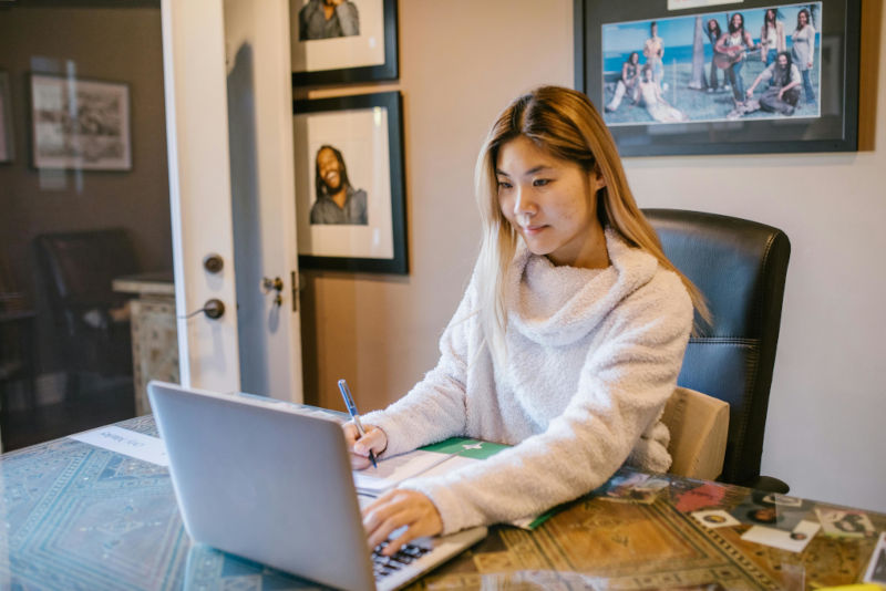 Person working at a dining table using a laptop and writing in a notebook, with framed photos on the wall behind them, suggesting focused remote or home office work.