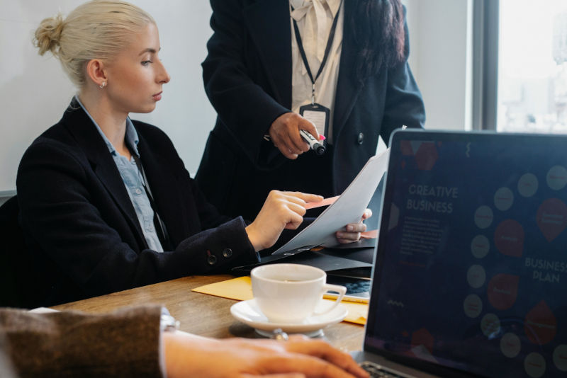 Team members reviewing a document together at a desk, with laptops open and a coffee cup nearby, showing focused collaboration in a busy work setting.