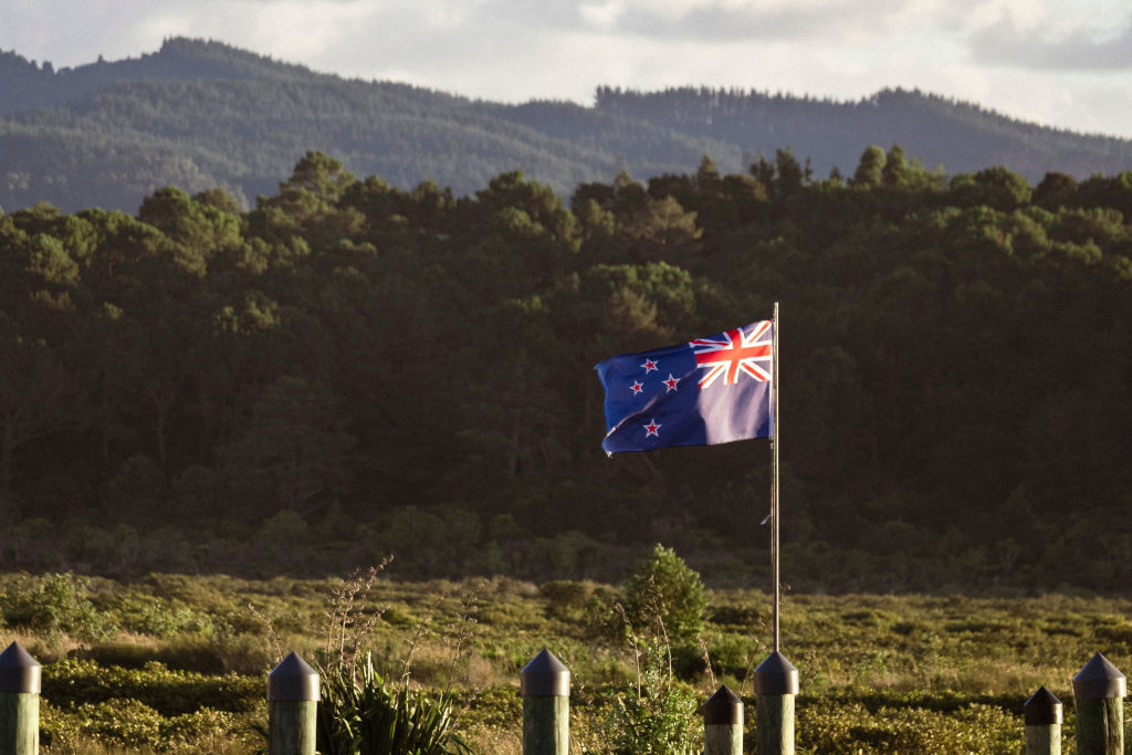A New Zealand flag waves against a backdrop of lush forests and mountains
