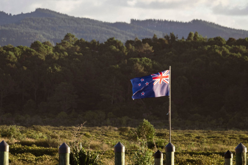 A New Zealand flag waves against a backdrop of lush forests and mountains