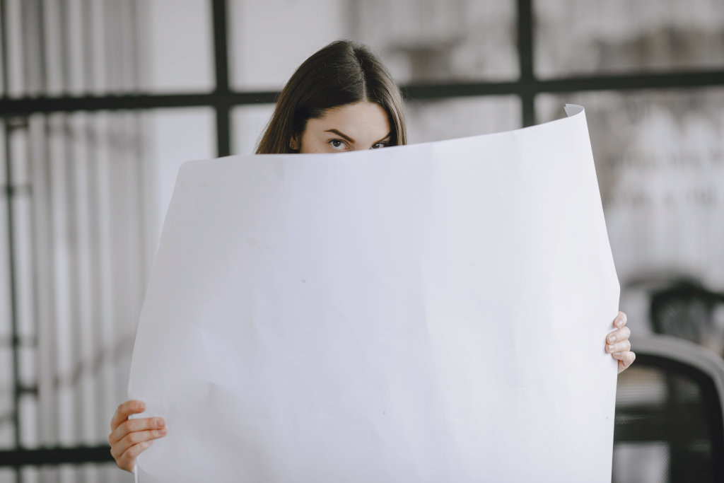 A woman partially hides behind a large white paper sheet indoors, looking directly at the camera