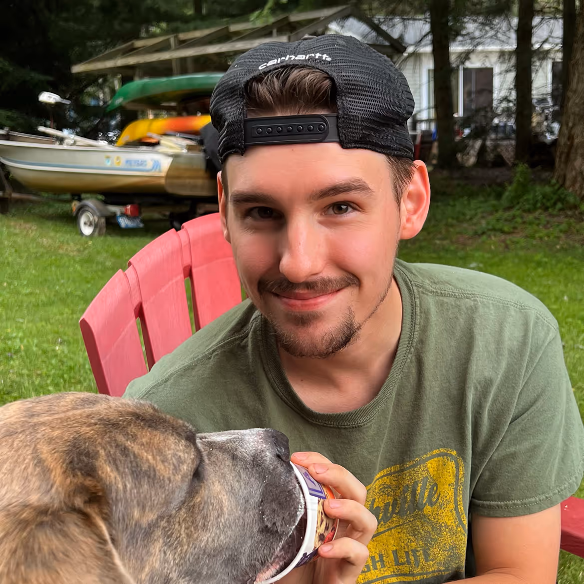 Man in backwards cap petting dog near boats and pink chair outdoors