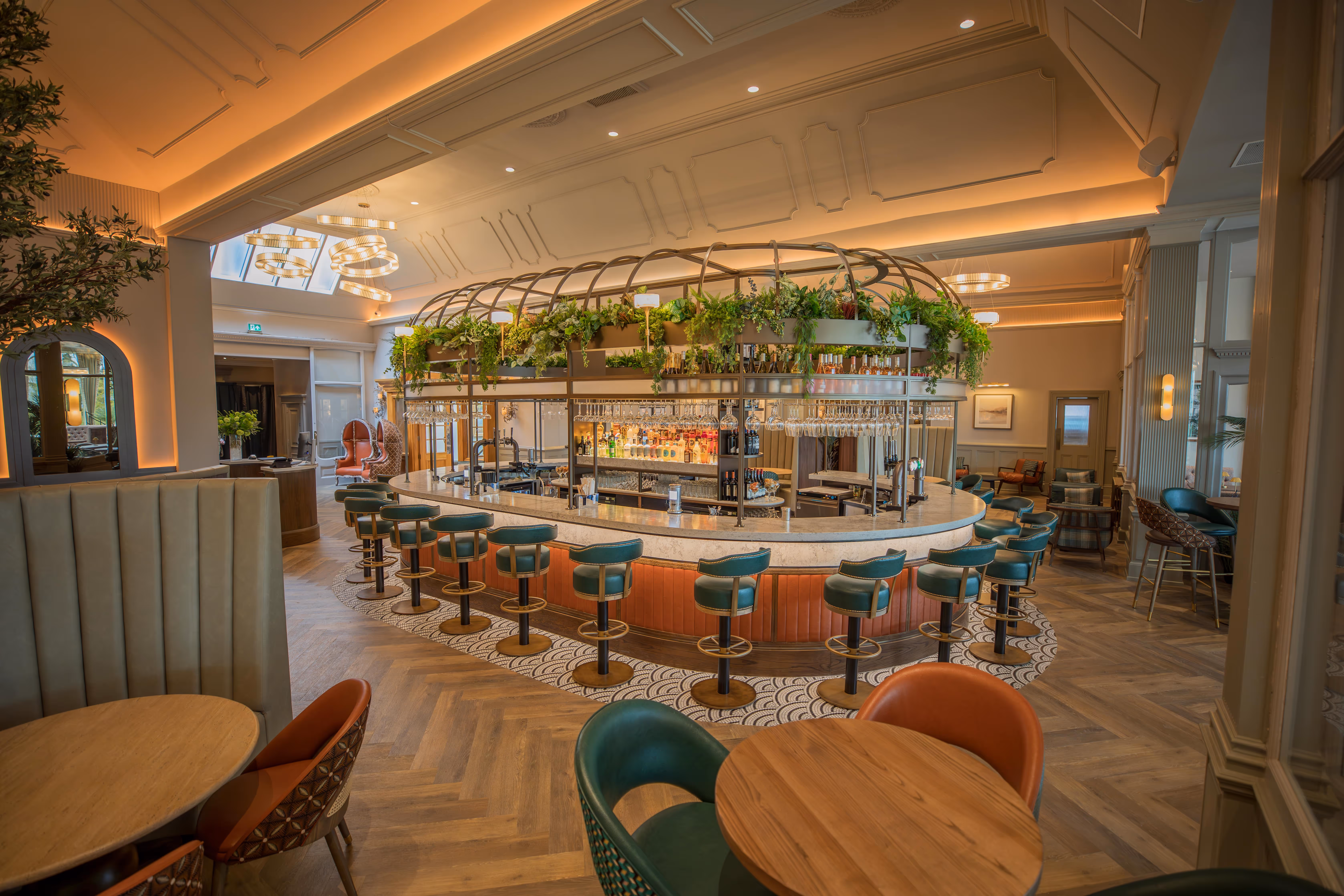Elegant modern bar with green bar stools, wooden tables, and greenery overhead under warm ambient lighting.