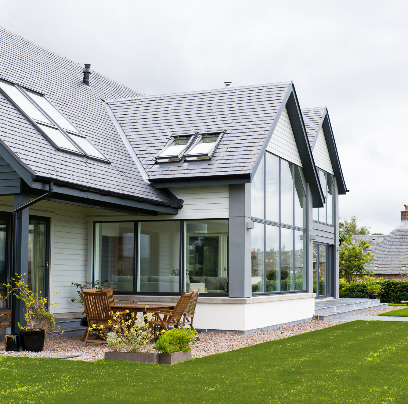 Modern house with large windows, gray tiled roof, and wooden patio furniture on a gravel area next to a green lawn.