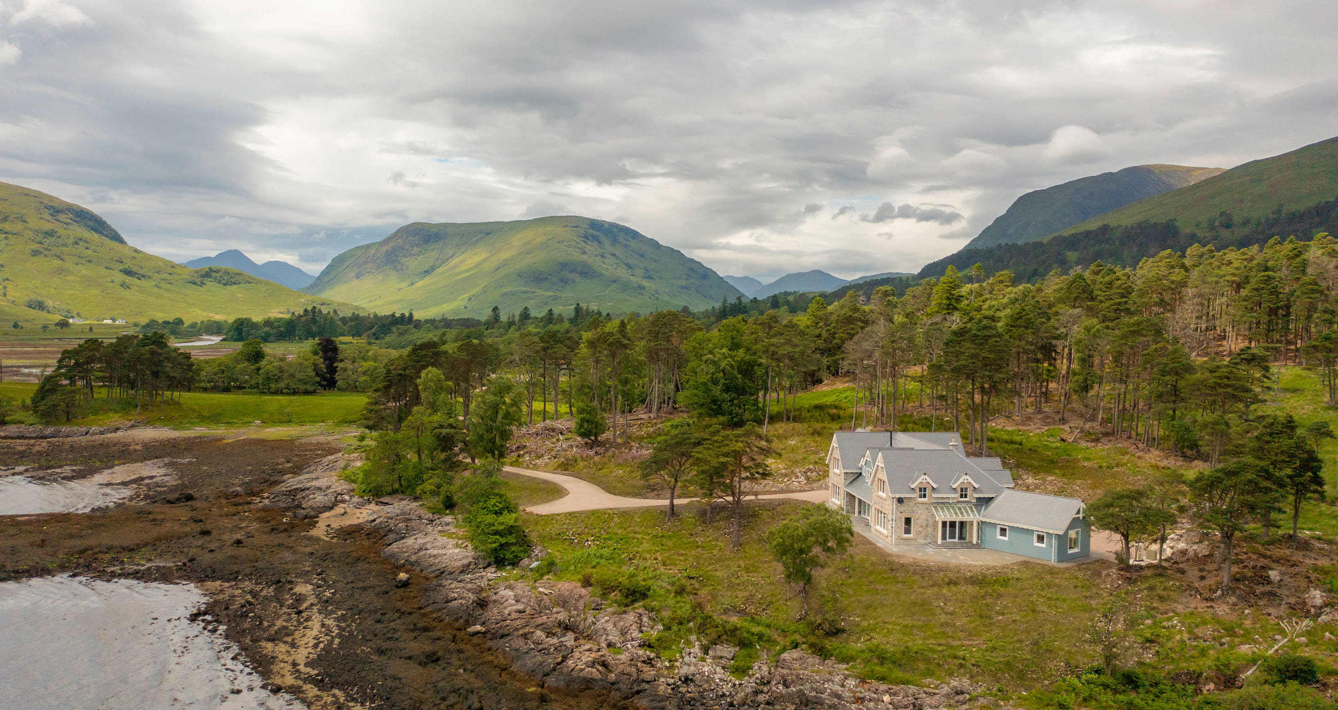 Stone and blue house near rocky shoreline with green hills and cloudy sky in the background.
