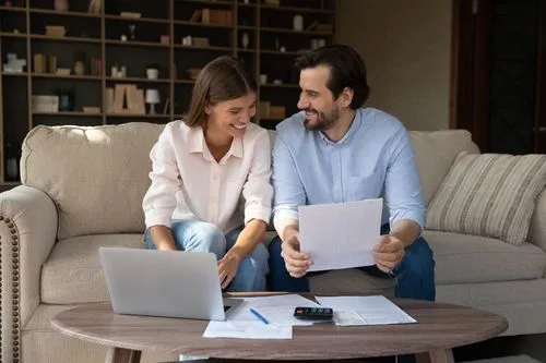 Smiling couple reviewing documents together on a couch with a laptop and papers on the table.