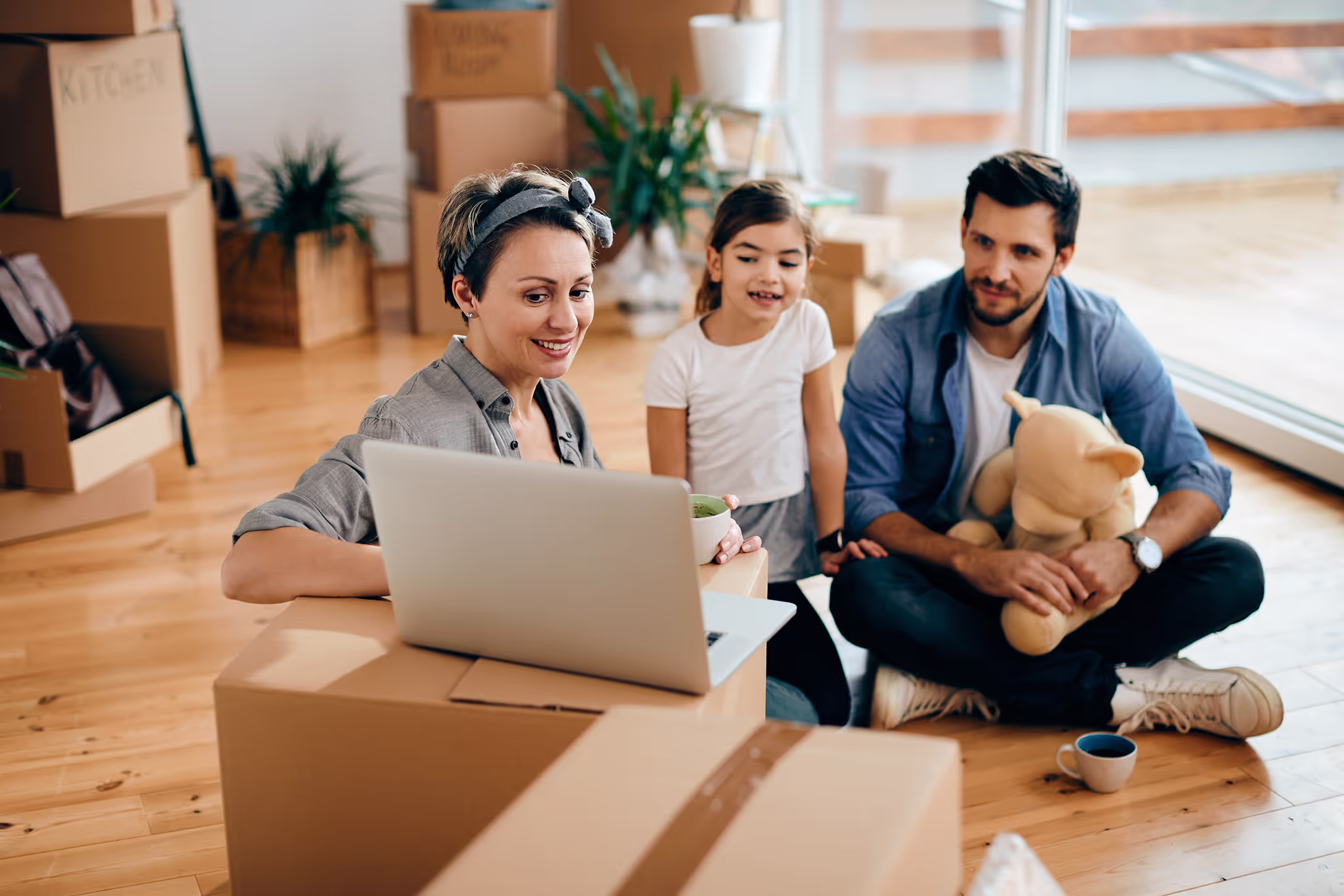 Family with young child sitting on floor surrounded by moving boxes, looking at a laptop.