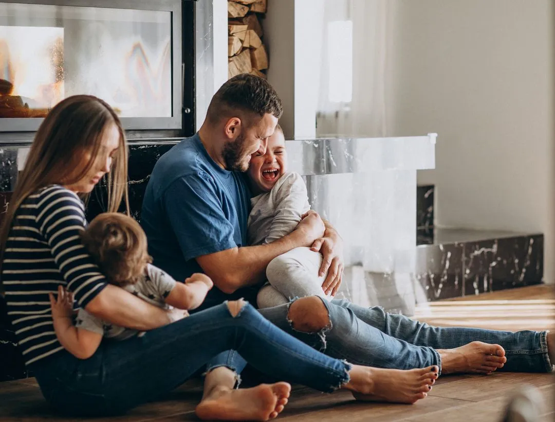 Family with two children sitting on floor, playing and laughing near a fireplace.