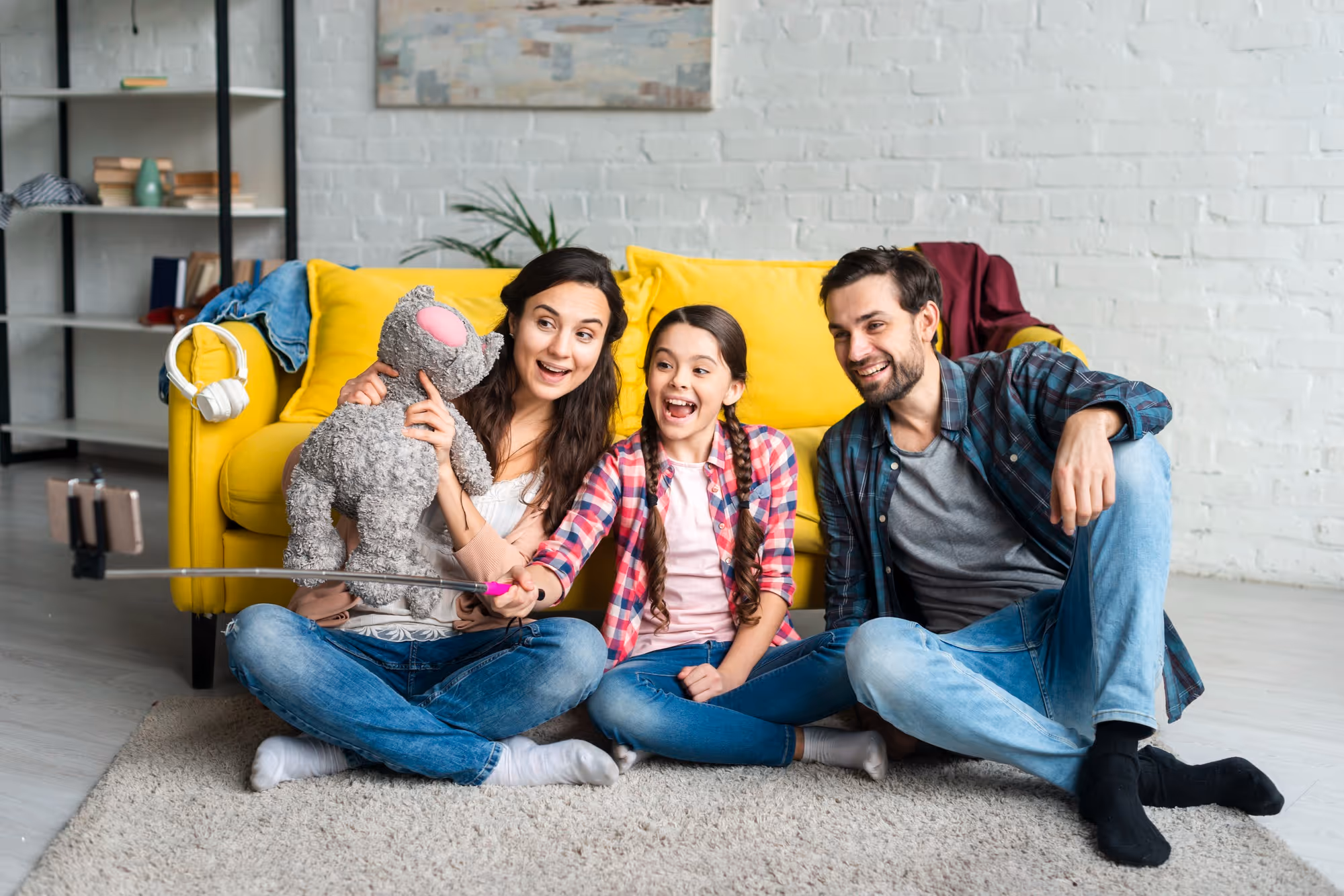 Happy family infornt of yellow sofa inside their home taking a selfie with gray teddy bear.