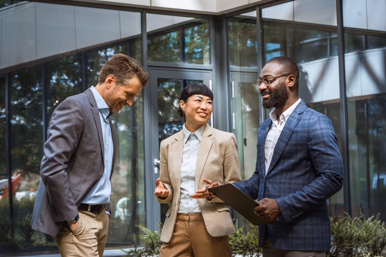Three business professionals smiling and talking outside a modern office building with glass walls.