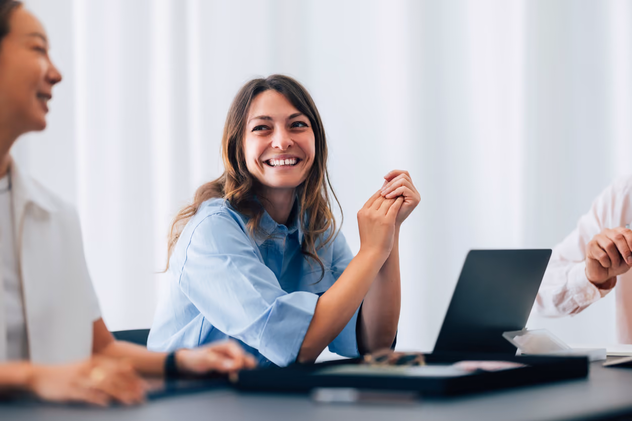 Young woman smiling and clasping hands at a meeting with two others and a laptop on the table.