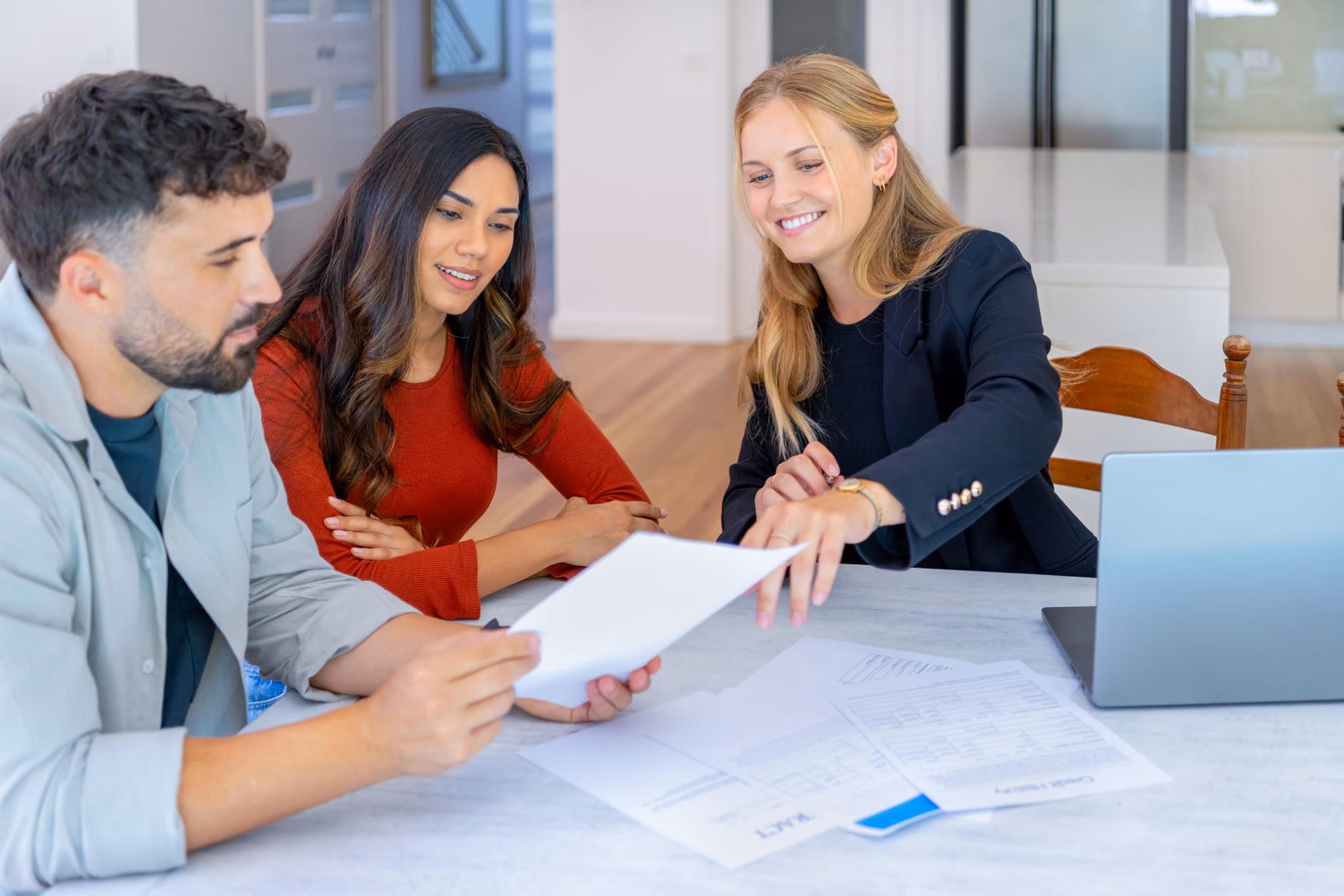 Three people reviewing documents at a table with a laptop in a bright office setting.