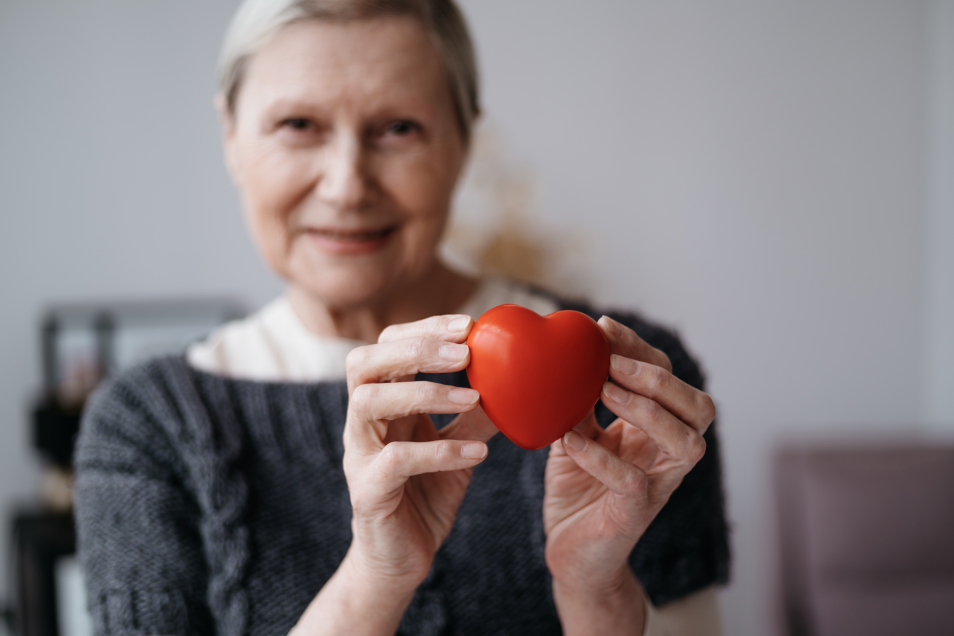 Une femme âgée souriante tient un cœur rouge en plastique devant elle.