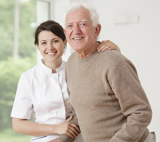Une aide-soignante souriante posant la main sur l'épaule d'un homme âgé souriant à l'intérieur d'une pièce lumineuse.