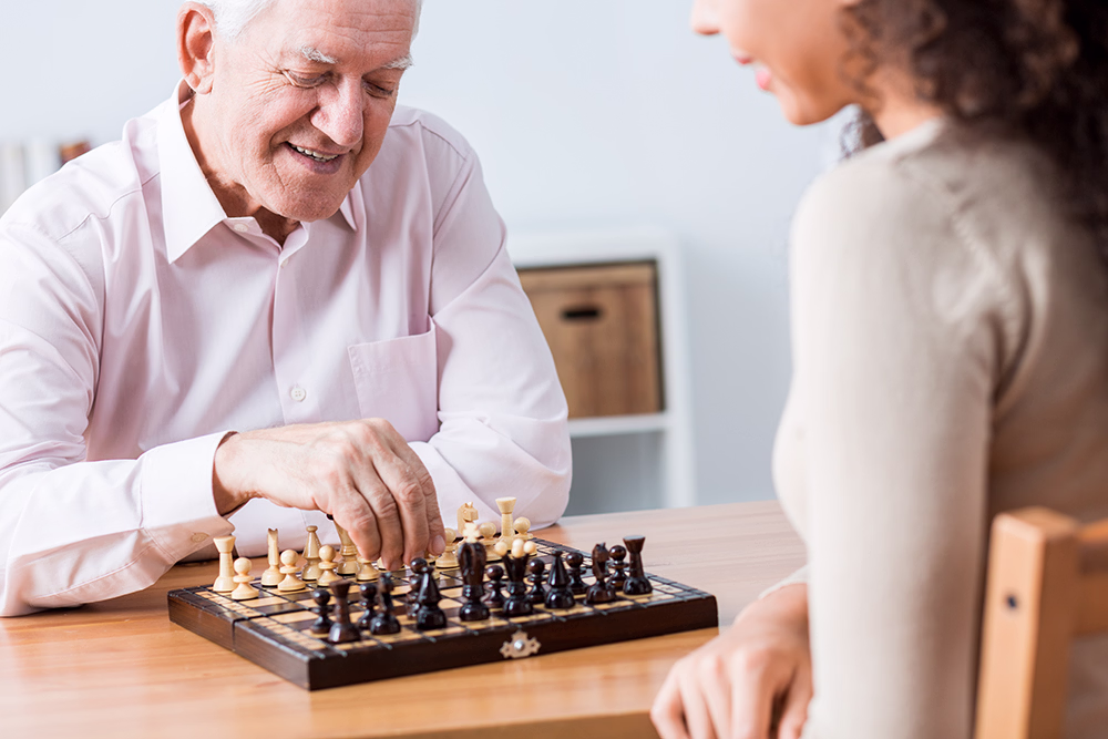 Un homme âgé souriant joue aux échecs avec une femme aux cheveux bouclés, assis à une table en bois.