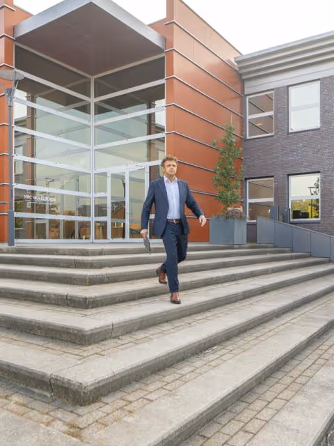 Man in een donker pak loopt de trappen af van een modern gebouw met grote glazen ramen en oranje accenten.