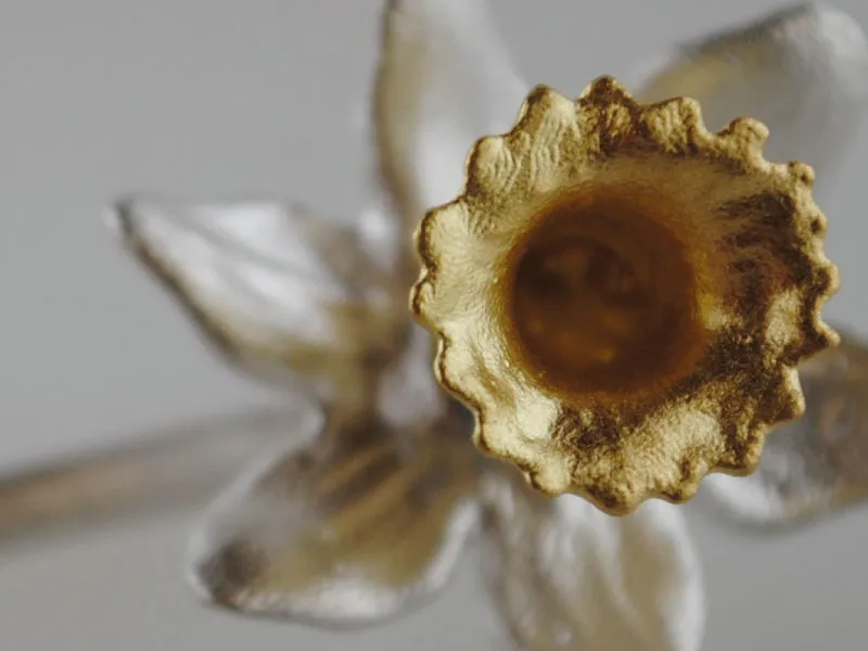 Close-up of a metallic gold daffodil flower sculpture with a blurred silver flower in the background.