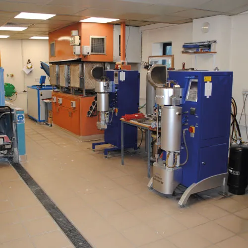 Industrial workshop with two blue and silver metal casting machines and an orange furnace in the background.