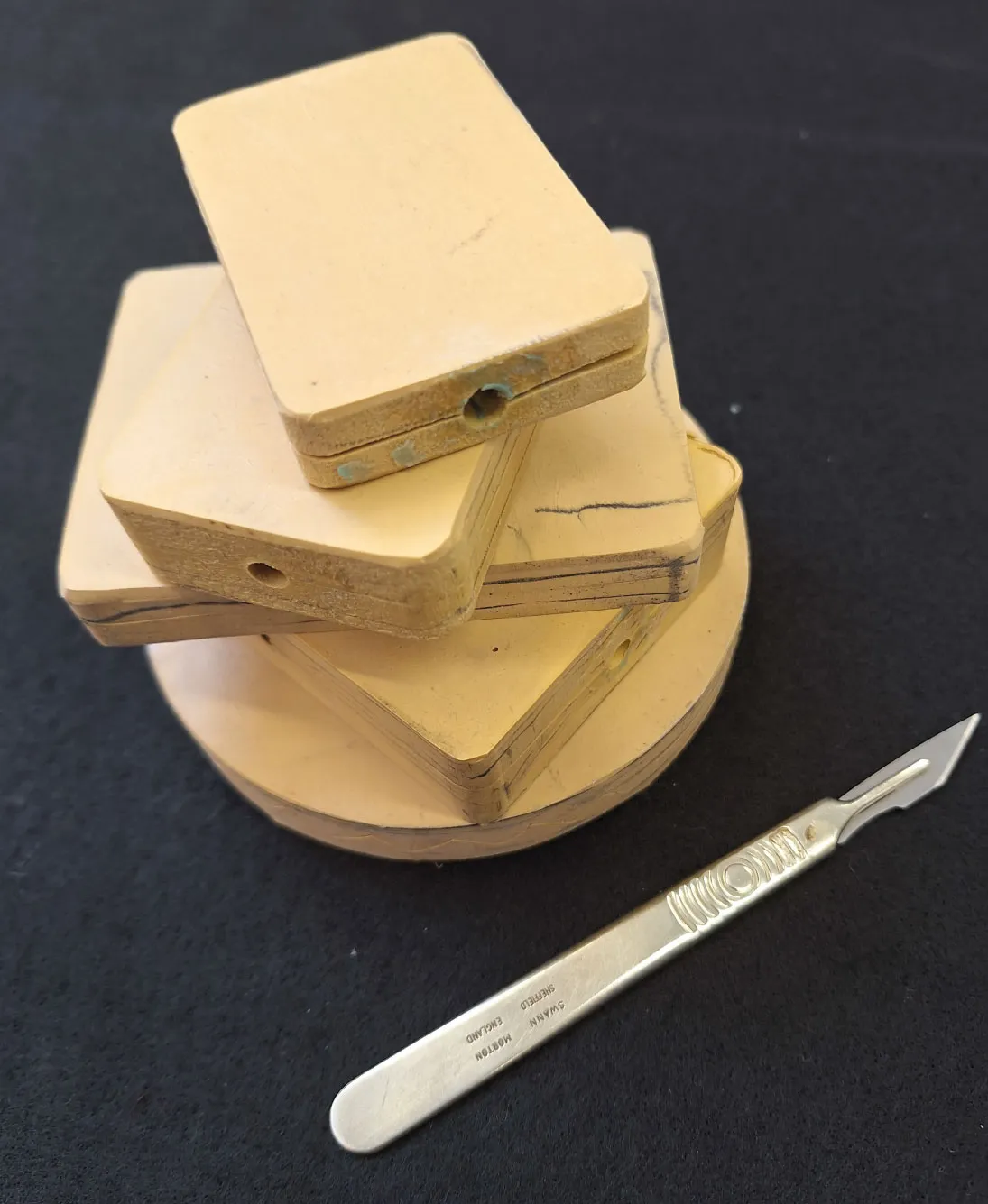 Stack of yellowish wooden blocks with holes, placed on a round wooden base, next to a metal scalpel with engravings.