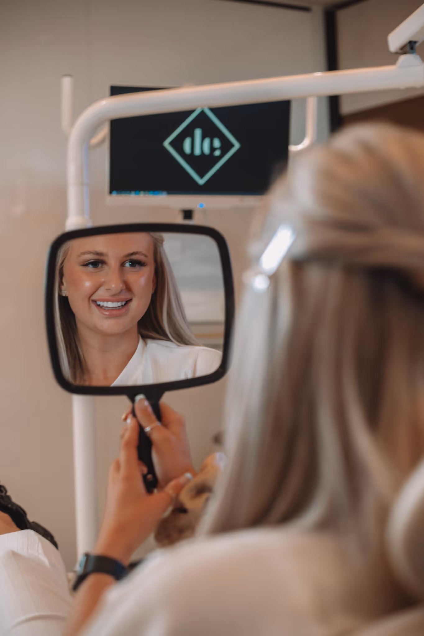 A woman in a white shirt smiling into a handheld mirror.