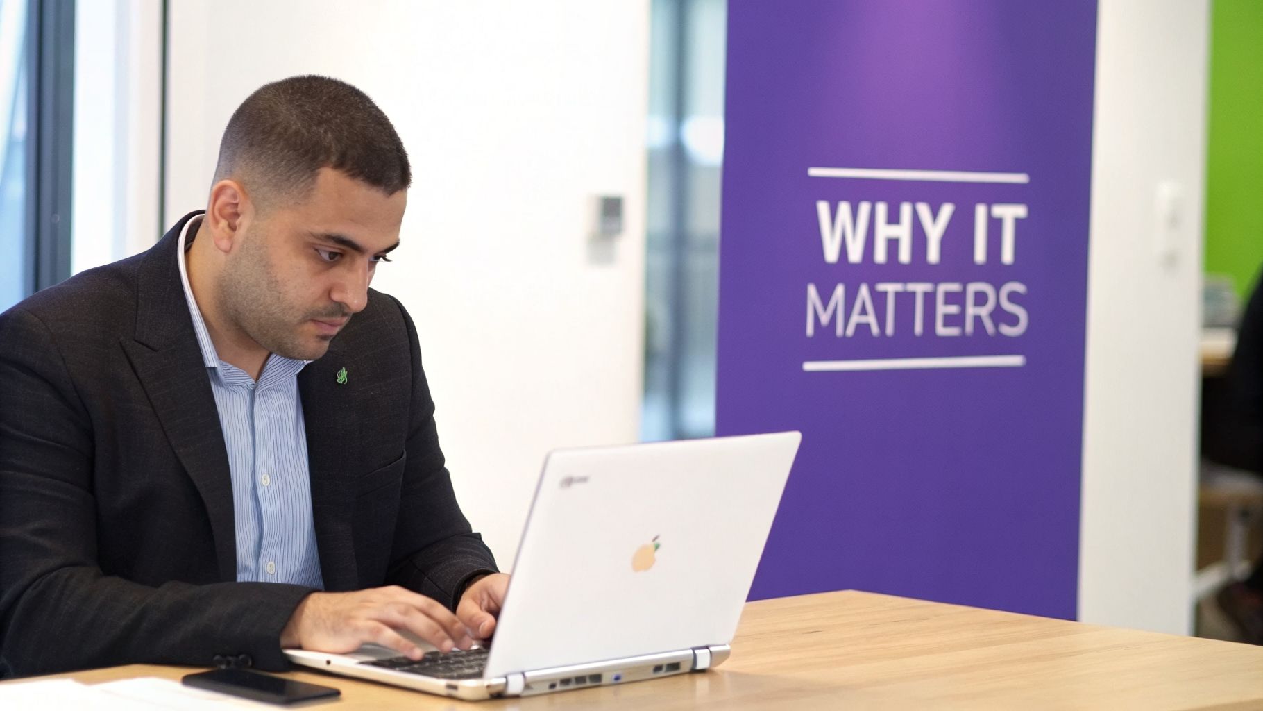 A professional man in a suit works on a laptop at a wooden table in an office.
